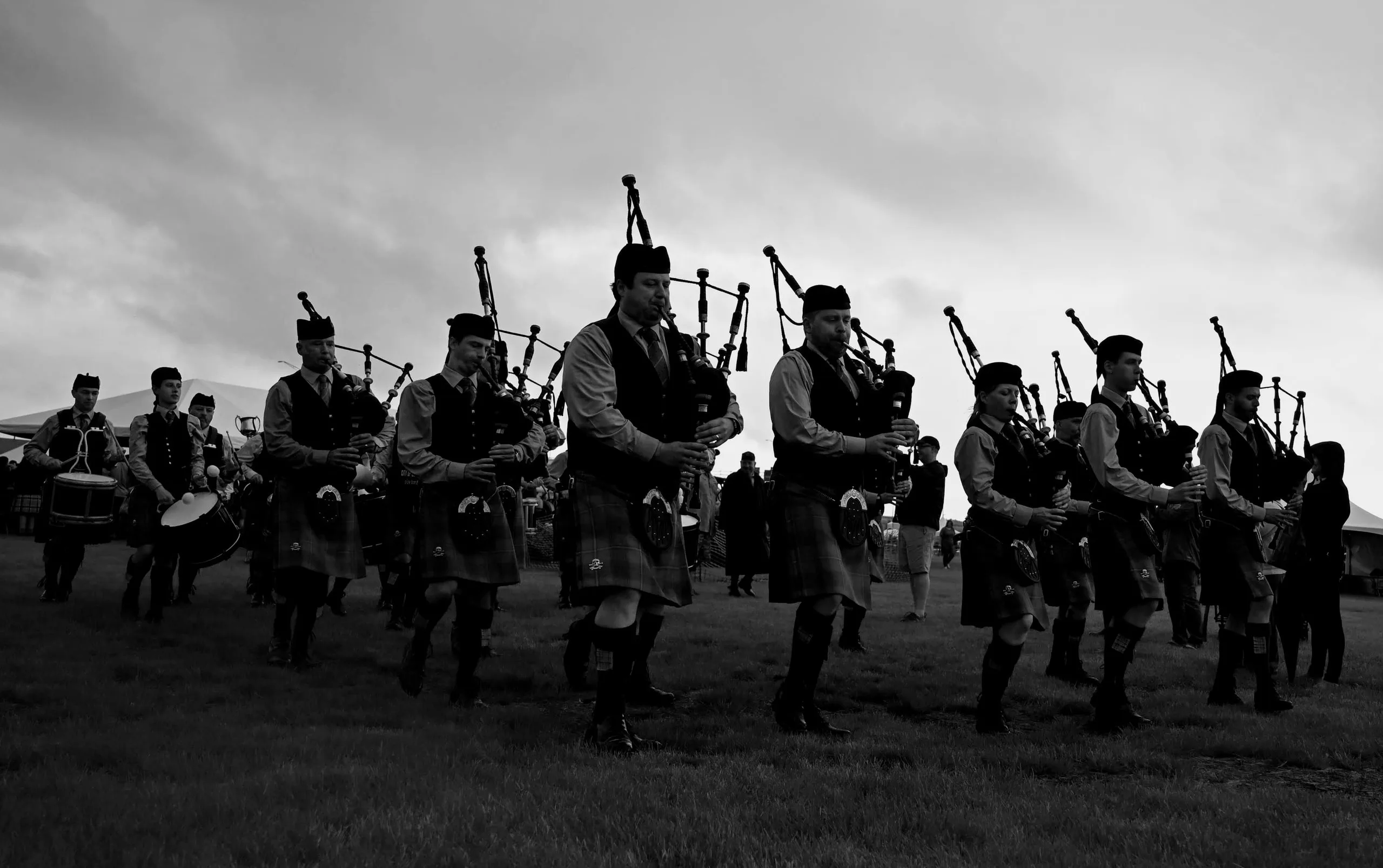 A black-and-white line of bagpipers and drummers marching across a field